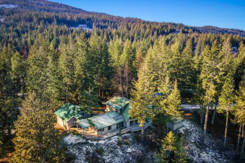 Aerial view of a house surrounded by tall evergreen trees and mountains under a clear blue sky.